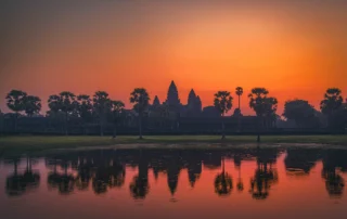 Angkor Wat tenpluaren eta palmondoen silueta urmaelean islatuta, zeru laranja biziaren azpian, Kanbodia
