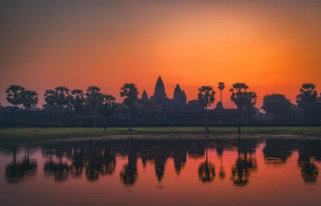 Angkor Wat tenpluaren eta palmondoen silueta urmaelean islatuta, zeru laranja biziaren azpian, Kanbodia