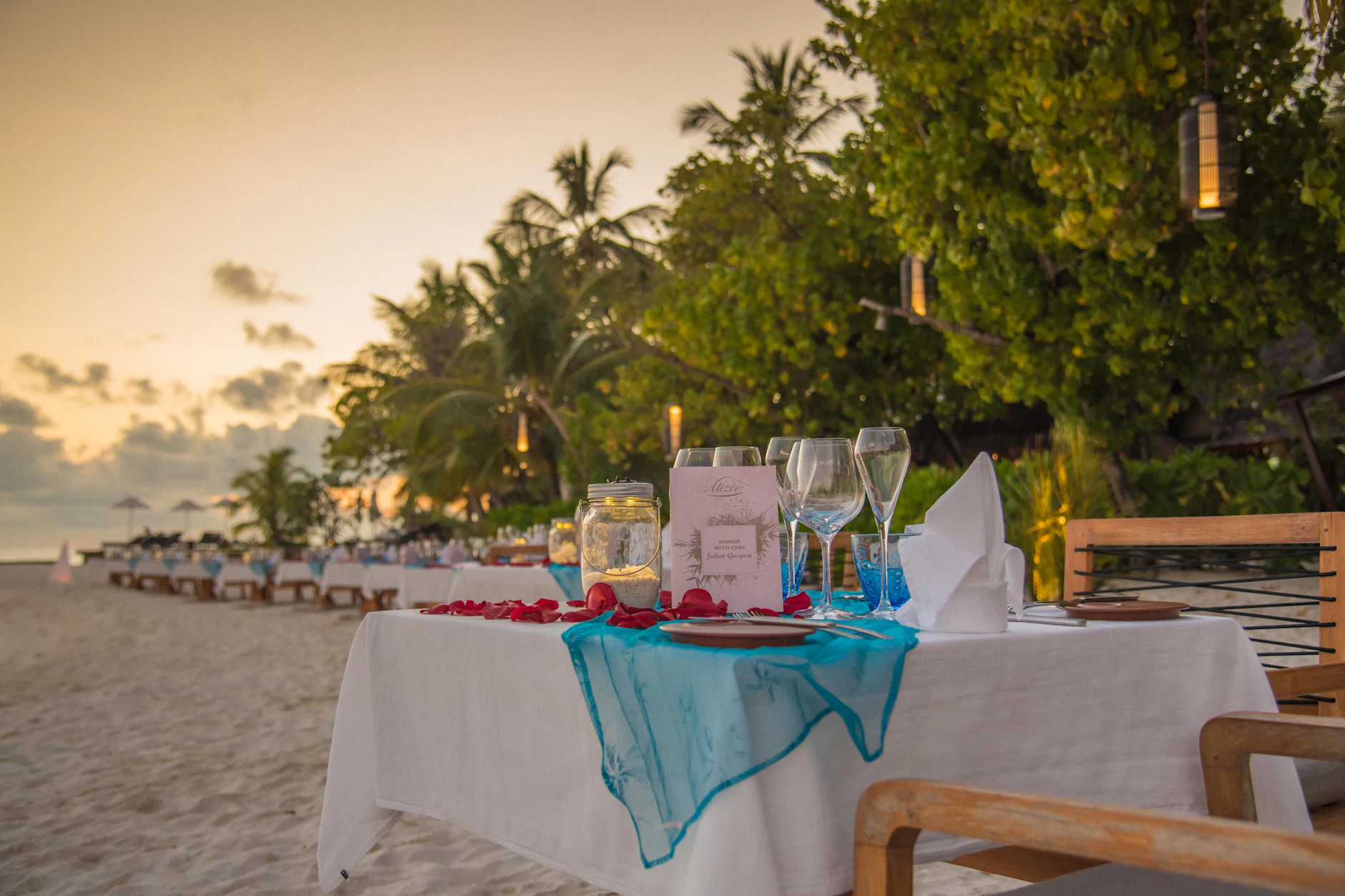 Mesa para cena romántica con pétalos y farolillos sobre la arena al atardecer en Maldivas