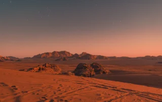 Wadi Rum basamortuko duna gorriak ilunabarrean izarrekin Wadi Rum basamortuko hare gorria eta mendiak ilunabarrean, lehen izarren azpian, Jordania