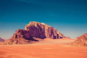 Gran macizo de roca rojiza que se alza sobre la arena anaranjada del desierto de Wadi Rum en Jordania