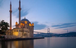 Mezquita de Ortaköy iluminada junto al Bósforo con el puente entre Europa y Asia al anochecer