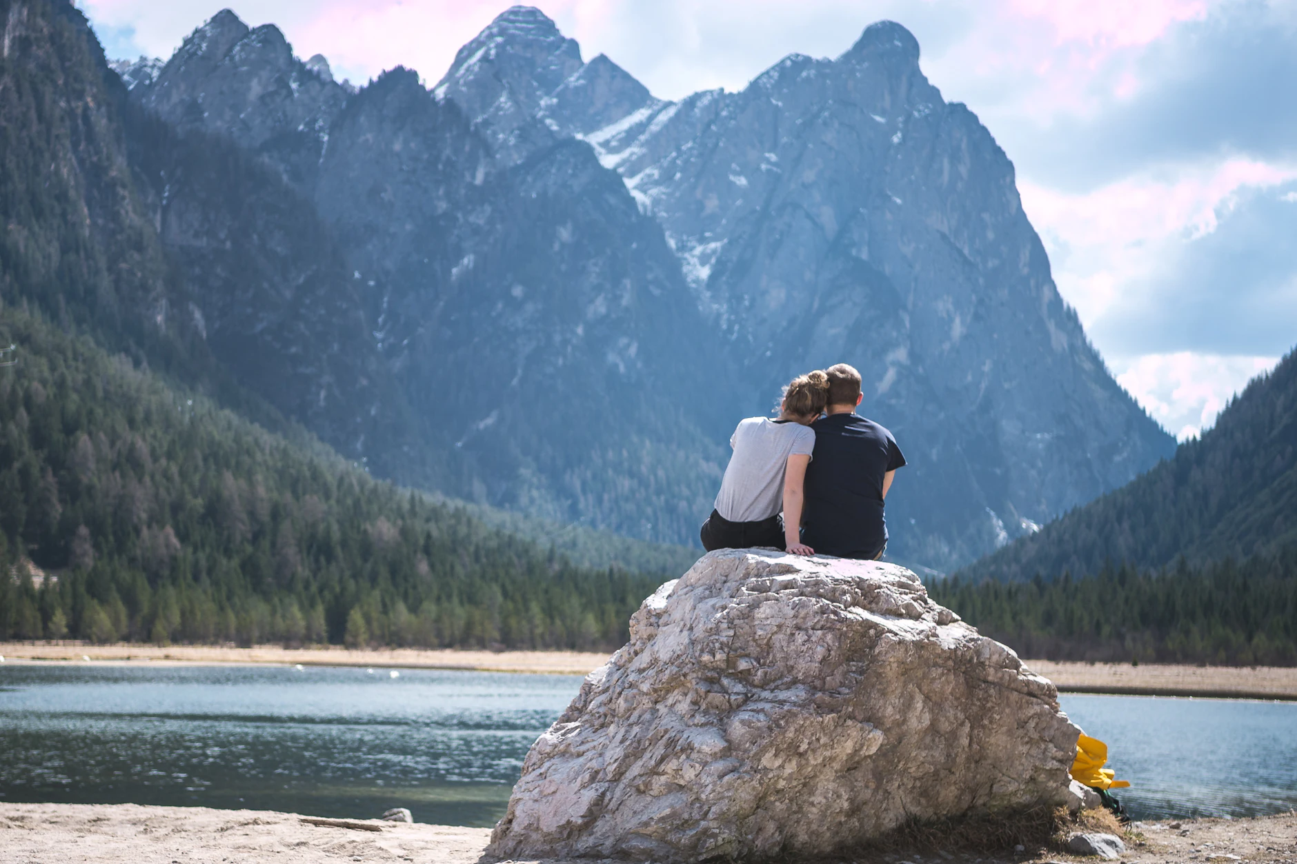 Pareja de espaldas sentada sobre una roca junto a un lago rodeado de montañas alpinas