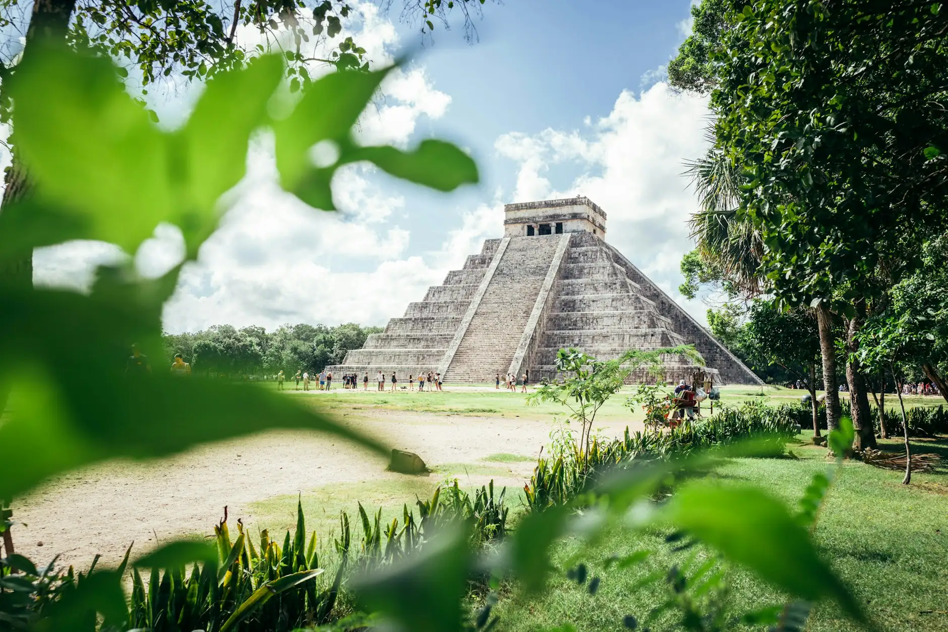 Pirámide de Kukulkán en Chichén Itzá entre la vegetación Pirámide de Kukulkán en Chichén Itzá, México, enmarcada por hojas verdes bajo un cielo azul